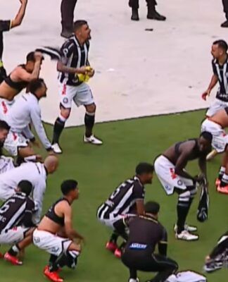 Jogadores e comissão técnica de um time de futebol, vestindo uniformes de listras verticais pretas e brancas com calções brancos, celebram intensamente em campo. Alguns atletas estão ajoelhados ou agachados no gramado, enquanto outros correm e agitam camisas no ar. Ao fundo, profissionais de imprensa com coletes e microfones acompanham a movimentação na borda do campo.