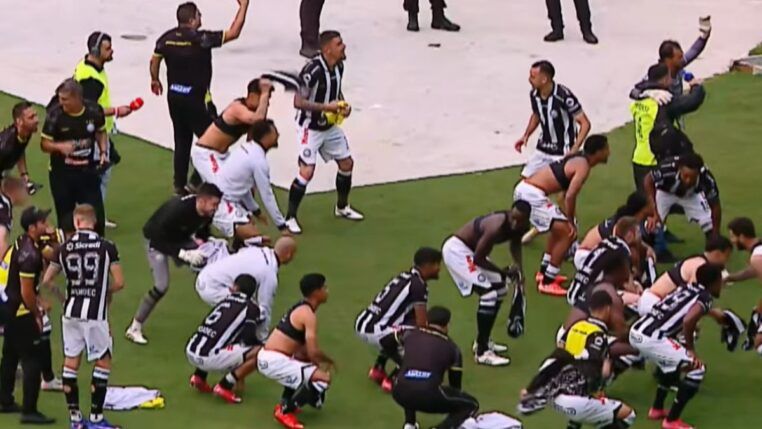 Jogadores e comissão técnica de um time de futebol, vestindo uniformes de listras verticais pretas e brancas com calções brancos, celebram intensamente em campo. Alguns atletas estão ajoelhados ou agachados no gramado, enquanto outros correm e agitam camisas no ar. Ao fundo, profissionais de imprensa com coletes e microfones acompanham a movimentação na borda do campo.