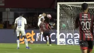 Registro de um lance de jogo de futebol à noite. Um jogador de uniforme branco salta para cabecear a bola em direção ao gol, enquanto um defensor de uniforme listrado em vermelho e preto tenta impedir a jogada. Em primeiro plano, as costas de outro jogador do mesmo time listrado, vestindo a camisa número 20, ocupam o lado direito da imagem. Ao fundo, vê-se a rede do gol e placas de publicidade no nível do gramado.