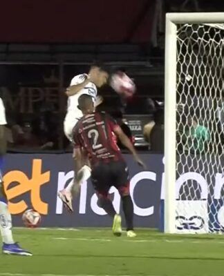Registro de um lance de jogo de futebol à noite. Um jogador de uniforme branco salta para cabecear a bola em direção ao gol, enquanto um defensor de uniforme listrado em vermelho e preto tenta impedir a jogada. Em primeiro plano, as costas de outro jogador do mesmo time listrado, vestindo a camisa número 20, ocupam o lado direito da imagem. Ao fundo, vê-se a rede do gol e placas de publicidade no nível do gramado.
