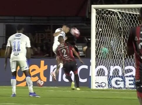 Registro de um lance de jogo de futebol à noite. Um jogador de uniforme branco salta para cabecear a bola em direção ao gol, enquanto um defensor de uniforme listrado em vermelho e preto tenta impedir a jogada. Em primeiro plano, as costas de outro jogador do mesmo time listrado, vestindo a camisa número 20, ocupam o lado direito da imagem. Ao fundo, vê-se a rede do gol e placas de publicidade no nível do gramado.