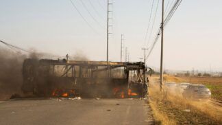 A carcaça de um ônibus incendiado ocupa o lado esquerdo de uma estrada asfaltada. Chamas e fumaça escura ainda são visíveis no interior do veículo destruído. À direita, carros de passeio trafegam por um acostamento de terra batida, levantando poeira. O cenário é cercado por vegetação seca e postes de energia altos sob um céu claro e ensolarado.