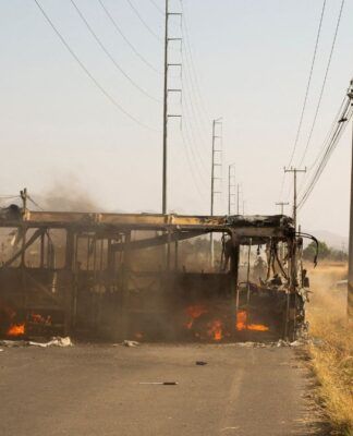 A carcaça de um ônibus incendiado ocupa o lado esquerdo de uma estrada asfaltada. Chamas e fumaça escura ainda são visíveis no interior do veículo destruído. À direita, carros de passeio trafegam por um acostamento de terra batida, levantando poeira. O cenário é cercado por vegetação seca e postes de energia altos sob um céu claro e ensolarado.