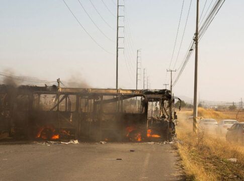 A carcaça de um ônibus incendiado ocupa o lado esquerdo de uma estrada asfaltada. Chamas e fumaça escura ainda são visíveis no interior do veículo destruído. À direita, carros de passeio trafegam por um acostamento de terra batida, levantando poeira. O cenário é cercado por vegetação seca e postes de energia altos sob um céu claro e ensolarado.