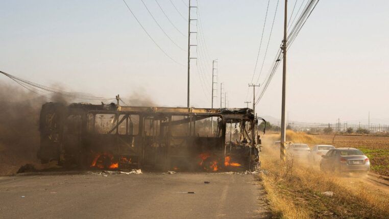 A carcaça de um ônibus incendiado ocupa o lado esquerdo de uma estrada asfaltada. Chamas e fumaça escura ainda são visíveis no interior do veículo destruído. À direita, carros de passeio trafegam por um acostamento de terra batida, levantando poeira. O cenário é cercado por vegetação seca e postes de energia altos sob um céu claro e ensolarado.