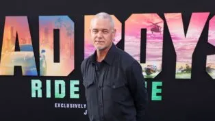 O ator Eric Dane, um homem de pele clara e cabelos curtos grisalhos, posa para uma foto em um tapete vermelho. Ele veste uma camisa social azul-marinho de botões. Ao fundo, há um painel preto com letras grandes coloridas que formam parte do título do filme "Bad Boys: Ride or Die", exibindo cenas de ação dentro das letras.