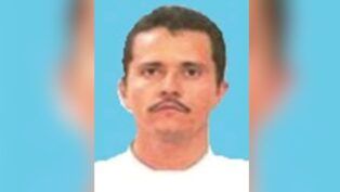 Uma foto de identificação de um homem de meia-idade com cabelos castanhos curtos e bigode. Ele está posicionado de frente contra um fundo azul sólido e veste o que parece ser uma camiseta branca. A imagem apresenta uma qualidade de resolução baixa e está levemente desfocada nas bordas laterais.