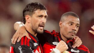 Dois jogadores de futebol do Flamengo estão em um momento de interação no campo. O jogador à esquerda, com barba e cabelo curto, está com o braço sobre o ombro do companheiro e parece falar algo com uma expressão intensa. O jogador à direita, com cabelo raspado, mantém uma expressão mais contida e séria. Ambos vestem a camisa rubro-negra (listras horizontais pretas e vermelhas) com o patrocínio "BRB" visível. O fundo está desfocado em tons de vermelho, sugerindo a presença da torcida no estádio.