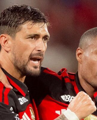 Dois jogadores de futebol do Flamengo estão em um momento de interação no campo. O jogador à esquerda, com barba e cabelo curto, está com o braço sobre o ombro do companheiro e parece falar algo com uma expressão intensa. O jogador à direita, com cabelo raspado, mantém uma expressão mais contida e séria. Ambos vestem a camisa rubro-negra (listras horizontais pretas e vermelhas) com o patrocínio 