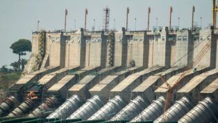 Vista externa da Usina Hidrelétrica de Belo Monte, mostrando a imensa estrutura de concreto da barragem. Na base, destacam-se os grandes condutos forçados cilíndricos e prateados que levam a água para as turbinas. No topo da estrutura, há diversas torres metálicas laranjas alinhadas, e ao fundo, à esquerda, uma área verde com árvores sob um céu claro.