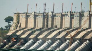 Vista externa da Usina Hidrelétrica de Belo Monte, mostrando a imensa estrutura de concreto da barragem. Na base, destacam-se os grandes condutos forçados cilíndricos e prateados que levam a água para as turbinas. No topo da estrutura, há diversas torres metálicas laranjas alinhadas, e ao fundo, à esquerda, uma área verde com árvores sob um céu claro.