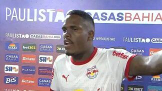 Um jogador de futebol negro, vestindo a camisa branca do Red Bull Bragantino, concede uma entrevista após uma partida. Ele está de perfil parcial, com a boca aberta como se estivesse falando e o braço esquerdo estendido. Ao fundo, há um painel de patrocinadores com as marcas "Paulistão Casas Bahia 2026", "Assaí", "Betano", entre outras. Dois microfones com protetores de espuma azuis aparecem na parte inferior da imagem.