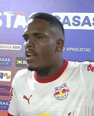 Um jogador de futebol negro, vestindo a camisa branca do Red Bull Bragantino, concede uma entrevista após uma partida. Ele está de perfil parcial, com a boca aberta como se estivesse falando e o braço esquerdo estendido. Ao fundo, há um painel de patrocinadores com as marcas 