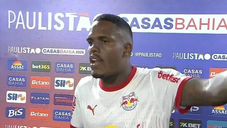 Um jogador de futebol negro, vestindo a camisa branca do Red Bull Bragantino, concede uma entrevista após uma partida. Ele está de perfil parcial, com a boca aberta como se estivesse falando e o braço esquerdo estendido. Ao fundo, há um painel de patrocinadores com as marcas "Paulistão Casas Bahia 2026", "Assaí", "Betano", entre outras. Dois microfones com protetores de espuma azuis aparecem na parte inferior da imagem.