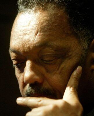 Close-up do rosto de um homem negro de cabelos grisalhos e bigode. Ele está com os olhos baixos, expressão séria e pensativa, com o dedo indicador encostado no queixo. O fundo está escuro e a iluminação foca em seu rosto.