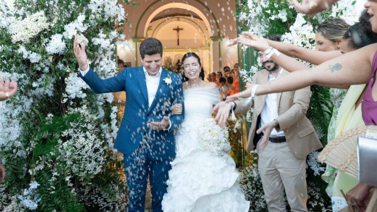 O casal de políticos João Campos e Tabata Amaral celebra seu casamento saindo de uma pequena igreja. Ambos estão sorridentes enquanto convidados jogam pétalas brancas sobre eles. João veste um terno azul-claro com uma flor branca na lapela, e Tabata usa um vestido de noiva branco ombro a ombro com camadas de pétalas de tecido na saia, segurando um buquê de flores brancas. A entrada da igreja é decorada com abundantes flores brancas e folhagens verdes.