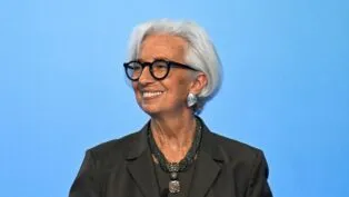 Uma mulher de cabelos brancos curtos e óculos de armação preta sorri para o lado. Ela veste um blazer escuro, um colar de várias voltas com um pingente quadrado e brincos prateados grandes. O fundo é de um azul claro uniforme.