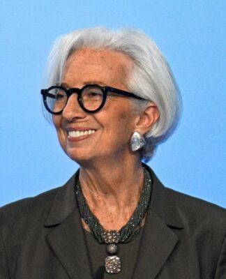 Uma mulher de cabelos brancos curtos e óculos de armação preta sorri para o lado. Ela veste um blazer escuro, um colar de várias voltas com um pingente quadrado e brincos prateados grandes. O fundo é de um azul claro uniforme.