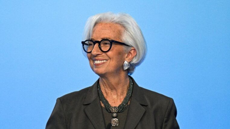 Uma mulher de cabelos brancos curtos e óculos de armação preta sorri para o lado. Ela veste um blazer escuro, um colar de várias voltas com um pingente quadrado e brincos prateados grandes. O fundo é de um azul claro uniforme.