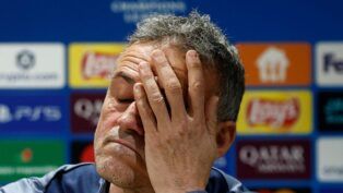 Um homem de pele clara e cabelos curtos grisalhos aparece em um plano fechado, com os olhos fechados e a mão cobrindo parte do rosto em um gesto de cansaço ou frustração. Ele veste uma peça de roupa azul. Ao fundo, há um painel desfocado com logotipos de patrocinadores e competições esportivas.