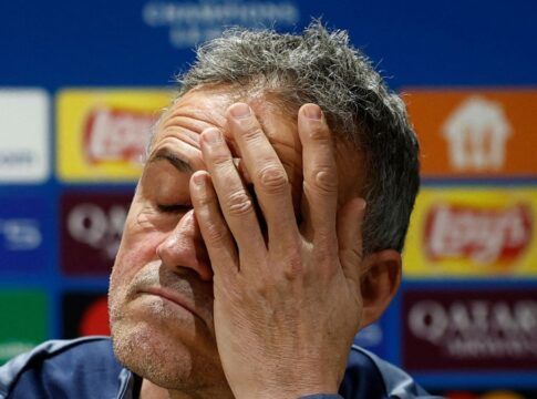 Um homem de pele clara e cabelos curtos grisalhos aparece em um plano fechado, com os olhos fechados e a mão cobrindo parte do rosto em um gesto de cansaço ou frustração. Ele veste uma peça de roupa azul. Ao fundo, há um painel desfocado com logotipos de patrocinadores e competições esportivas.