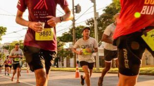 Vários corredores participam de uma prova de rua durante o dia. Em destaque no centro, uma jovem veste uma camiseta cinza com o logotipo "Meia de Curita", boné escuro e calções pretos, mantendo o foco na corrida. À esquerda, em primeiro plano, um homem veste uma camiseta vinho com a inscrição "Corrida da Ponte" e o número de peito 3802, segurando um copo de água. Outros corredores são visíveis ao fundo em uma avenida arborizada, sob a luz do sol. Postes de energia e fiação elétrica cruzam o céu ao fundo.