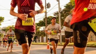 Vários corredores participam de uma prova de rua durante o dia. Em destaque no centro, uma jovem veste uma camiseta cinza com o logotipo "Meia de Curita", boné escuro e calções pretos, mantendo o foco na corrida. À esquerda, em primeiro plano, um homem veste uma camiseta vinho com a inscrição "Corrida da Ponte" e o número de peito 3802, segurando um copo de água. Outros corredores são visíveis ao fundo em uma avenida arborizada, sob a luz do sol. Postes de energia e fiação elétrica cruzam o céu ao fundo.