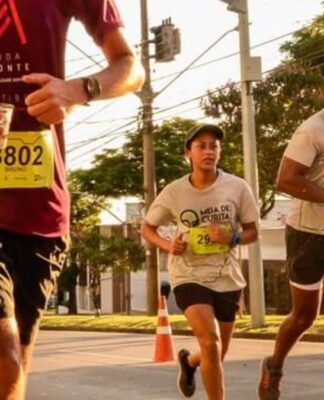Vários corredores participam de uma prova de rua durante o dia. Em destaque no centro, uma jovem veste uma camiseta cinza com o logotipo 