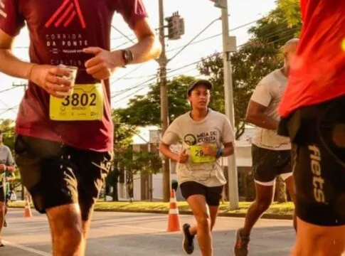 Vários corredores participam de uma prova de rua durante o dia. Em destaque no centro, uma jovem veste uma camiseta cinza com o logotipo 