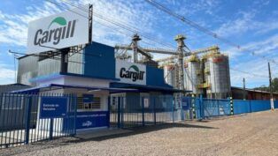 Vista externa de uma unidade industrial da Cargill sob um céu azul com nuvens esparsas. Em primeiro plano, há uma guarita branca e azul com uma placa indicando "Entrada de Veículos". Um grande outdoor e a fachada do prédio principal exibem o logotipo da "Cargill" em verde e cinza. Ao fundo, estruturas metálicas altas e vários silos cilíndricos cinzas compõem o complexo industrial. O chão à frente é de terra batida e brita, delimitado por grades e portões metálicos azuis.