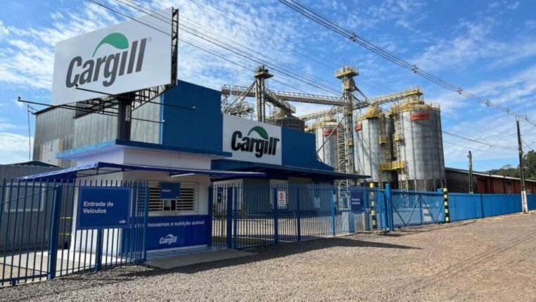 Vista externa de uma unidade industrial da Cargill sob um céu azul com nuvens esparsas. Em primeiro plano, há uma guarita branca e azul com uma placa indicando "Entrada de Veículos". Um grande outdoor e a fachada do prédio principal exibem o logotipo da "Cargill" em verde e cinza. Ao fundo, estruturas metálicas altas e vários silos cilíndricos cinzas compõem o complexo industrial. O chão à frente é de terra batida e brita, delimitado por grades e portões metálicos azuis.