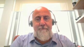 Um homem de pele clara e calvo, com uma barba grisalha bem aparada, olha diretamente para a câmera. Ele utiliza um headset preto (fone de ouvido com microfone acoplado) e veste uma camisa social azul clara. O fundo é composto por uma parede branca iluminada e parte de uma estante de madeira à direita.