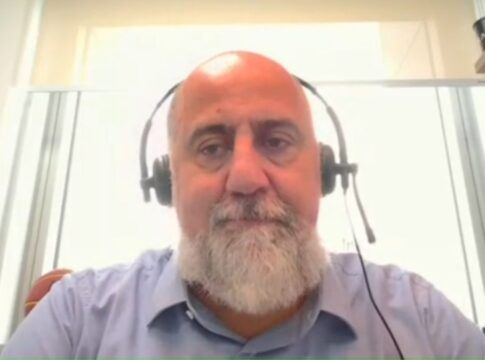 Um homem de pele clara e calvo, com uma barba grisalha bem aparada, olha diretamente para a câmera. Ele utiliza um headset preto (fone de ouvido com microfone acoplado) e veste uma camisa social azul clara. O fundo é composto por uma parede branca iluminada e parte de uma estante de madeira à direita.