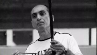 Fotografia em preto e branco de um homem de meia-idade, calvo no topo da cabeça, com expressão serena. Ele veste uma camiseta clara e segura um berimbau na posição vertical, com a mão esquerda no instrumento e a direita segurando a baqueta.