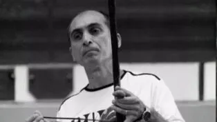 Fotografia em preto e branco de um homem de meia-idade, calvo no topo da cabeça, com expressão serena. Ele veste uma camiseta clara e segura um berimbau na posição vertical, com a mão esquerda no instrumento e a direita segurando a baqueta.