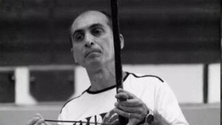 Fotografia em preto e branco de um homem de meia-idade, calvo no topo da cabeça, com expressão serena. Ele veste uma camiseta clara e segura um berimbau na posição vertical, com a mão esquerda no instrumento e a direita segurando a baqueta.