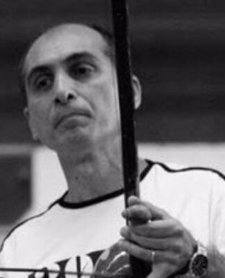 Fotografia em preto e branco de um homem de meia-idade, calvo no topo da cabeça, com expressão serena. Ele veste uma camiseta clara e segura um berimbau na posição vertical, com a mão esquerda no instrumento e a direita segurando a baqueta.