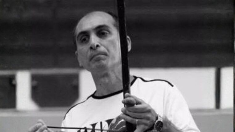 Fotografia em preto e branco de um homem de meia-idade, calvo no topo da cabeça, com expressão serena. Ele veste uma camiseta clara e segura um berimbau na posição vertical, com a mão esquerda no instrumento e a direita segurando a baqueta.