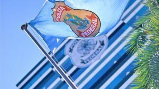 Bandeira azul clara da Polícia Federal hasteada e ondulando ao vento sob um céu azul. Ao fundo, de forma desfocada, vê-se a fachada de um prédio moderno com vidros espelhados e outro logotipo da instituição. À direita, parte das folhas de uma palmeira emoldura a cena.