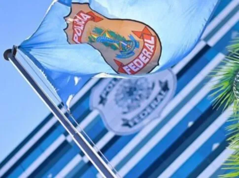 Bandeira azul clara da Polícia Federal hasteada e ondulando ao vento sob um céu azul. Ao fundo, de forma desfocada, vê-se a fachada de um prédio moderno com vidros espelhados e outro logotipo da instituição. À direita, parte das folhas de uma palmeira emoldura a cena.