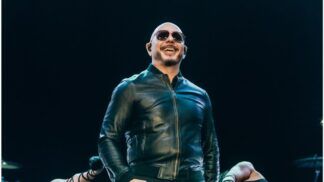 O cantor Pitbull, um homem calvo de pele clara, sorri enquanto se apresenta em um palco. Ele usa óculos de sol escuros, uma jaqueta de couro preta fechada e fones de ouvido de monitoramento (in-ear). O fundo é escuro, com luzes de palco focadas nele, e parte dos braços de dançarinas é visível na parte inferior da imagem.