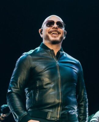 O cantor Pitbull, um homem calvo de pele clara, sorri enquanto se apresenta em um palco. Ele usa óculos de sol escuros, uma jaqueta de couro preta fechada e fones de ouvido de monitoramento (in-ear). O fundo é escuro, com luzes de palco focadas nele, e parte dos braços de dançarinas é visível na parte inferior da imagem.