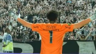 Um goleiro de costas, vestindo uma camisa laranja vibrante com o número 1 e o patrocínio "EZZE" em destaque, estende os braços em direção à arquibancada. Ele usa luvas brancas e cinzas. À sua frente, uma multidão de torcedores lota o estádio, muitos deles com os braços levantados em comemoração ou saudação. Uma faixa preta horizontal é visível na base da arquibancada, e um profissional de colete amarelo e azul aparece no canto inferior esquerdo.