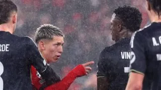 Sob chuva intensa, dois jogadores de futebol interagem no gramado durante uma partida noturna. O jogador à esquerda, com cabelo loiro curto e descolorido, veste um uniforme vermelho de mangas longas e aponta o dedo indicador em direção ao outro atleta. O jogador à direita, de costas para a câmera, veste um uniforme preto com o nome "VINI" e o número 7 visíveis. Outros dois jogadores com uniformes pretos aparecem parcialmente nas laterais da imagem. O fundo está desfocado, mostrando as luzes e as cores das arquibancadas do estádio.