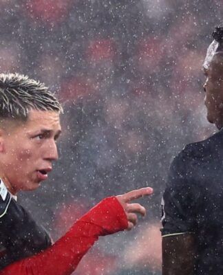Sob chuva intensa, dois jogadores de futebol interagem no gramado durante uma partida noturna. O jogador à esquerda, com cabelo loiro curto e descolorido, veste um uniforme vermelho de mangas longas e aponta o dedo indicador em direção ao outro atleta. O jogador à direita, de costas para a câmera, veste um uniforme preto com o nome 