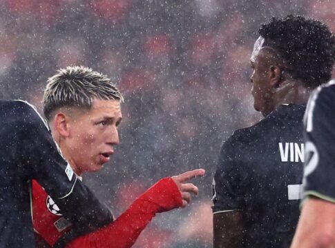 Sob chuva intensa, dois jogadores de futebol interagem no gramado durante uma partida noturna. O jogador à esquerda, com cabelo loiro curto e descolorido, veste um uniforme vermelho de mangas longas e aponta o dedo indicador em direção ao outro atleta. O jogador à direita, de costas para a câmera, veste um uniforme preto com o nome 