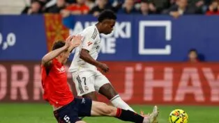 O jogador Vinícius Júnior, vestindo o uniforme branco do Real Madrid, está em movimento com a bola durante uma partida de futebol. Ele é confrontado por um adversário de uniforme vermelho e calção azul, que tenta desarmá-lo com um carrinho. Ao fundo, é possível ver a arquibancada com torcedores e placas de publicidade coloridas.