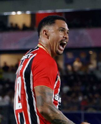 O jogador Luciano, do São Paulo FC, celebra intensamente em campo durante uma partida noturna. Ele está de perfil, com a boca aberta em um grito de comemoração e os punhos cerrados. Veste a camisa listrada em vermelho, branco e preto, calções brancos e meiões brancos. Ao fundo, as arquibancadas do estádio estão lotadas e iluminadas pelos refletores.