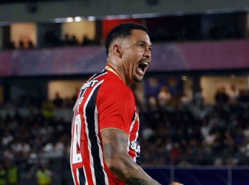 O jogador Luciano, do São Paulo FC, celebra intensamente em campo durante uma partida noturna. Ele está de perfil, com a boca aberta em um grito de comemoração e os punhos cerrados. Veste a camisa listrada em vermelho, branco e preto, calções brancos e meiões brancos. Ao fundo, as arquibancadas do estádio estão lotadas e iluminadas pelos refletores.