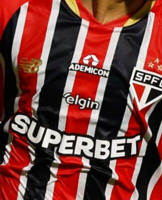 Close-up do tronco de um jogador vestindo a camisa listrada do São Paulo Futebol Clube, em vermelho, preto e branco. Diversos logotipos de patrocinadores estão visíveis no uniforme, incluindo o escudo do time no peito e marcas comerciais no centro e ombros.